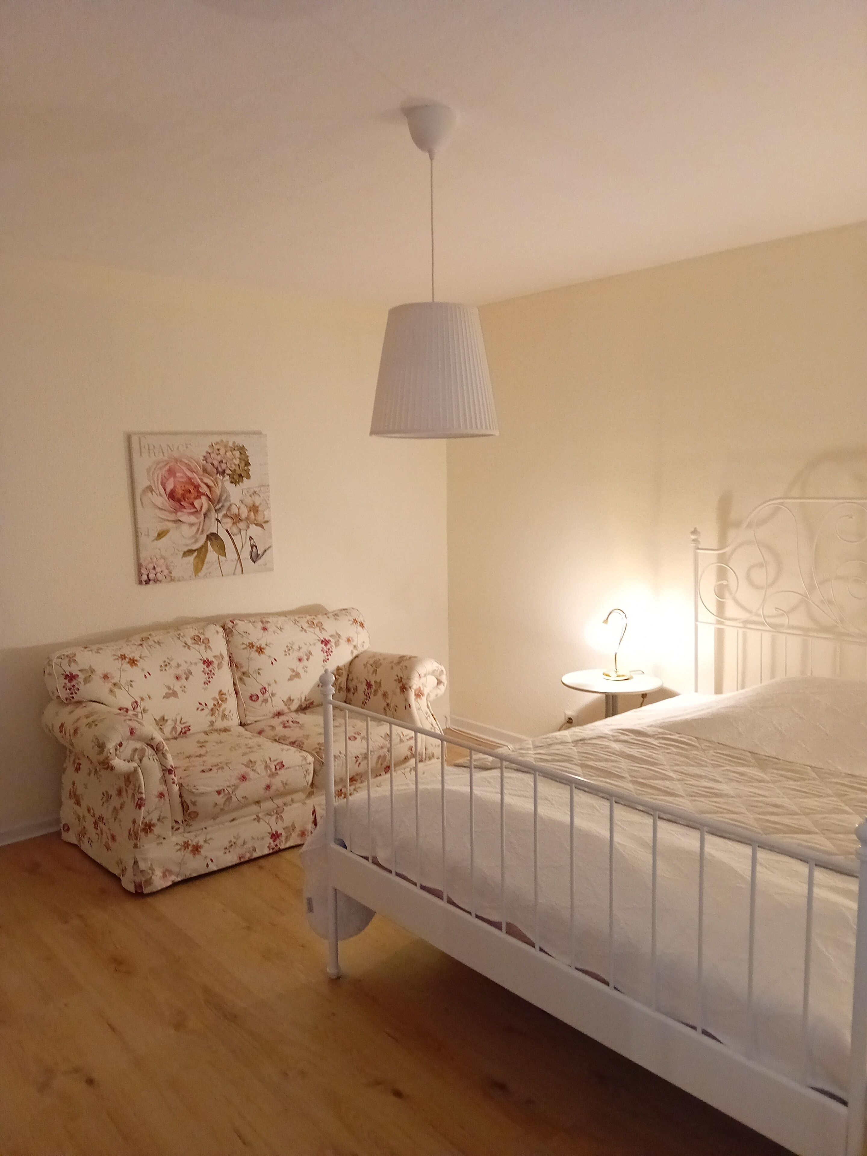 Un dormitori amb un llit blanc, un sofà floral, una làmpada penjant i un quadre a la paret.