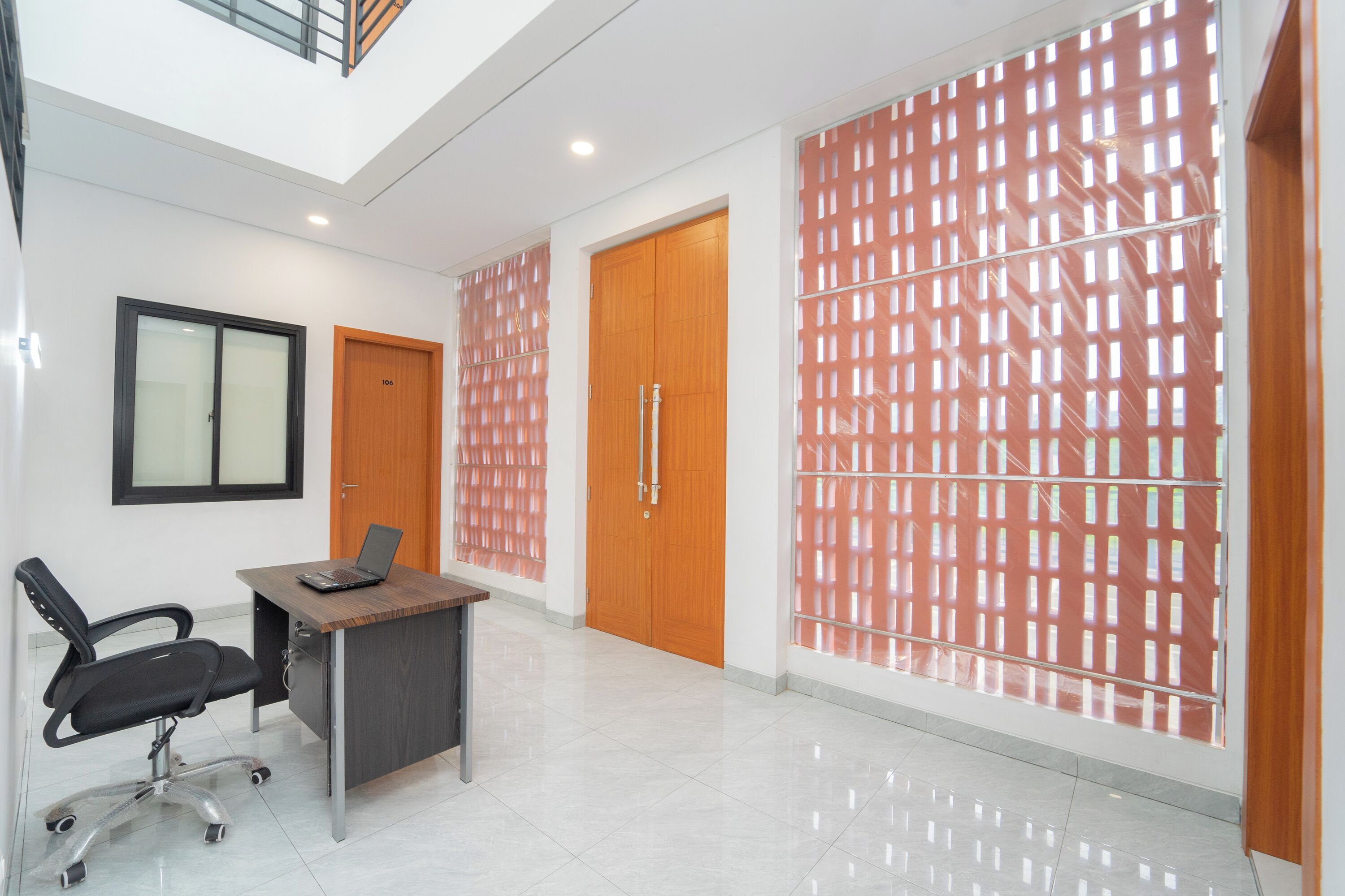 A modern office space with a desk, office chair, and a laptop. There are two wooden doors and a large window with a patterned screen.