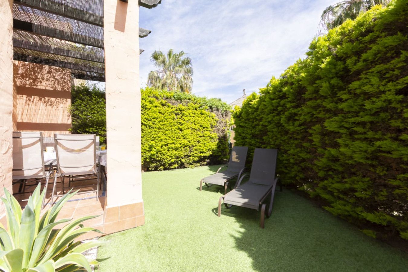 Eine Terrassenfläche mit zwei Liegestühlen und einem Tisch, umgeben von üppigem Grün und einem gepflegten Rasen.