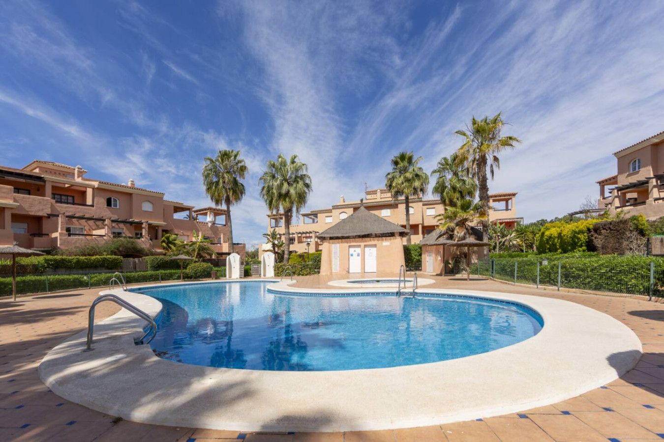 Una piscina de forma circular, rodeada por una solana empedrada, junto a un edificio con varios balcones y palmeras.