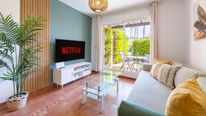 Ein Wohnzimmer mit einem Flachbildfernseher, der 'NETFLIX' zeigt, einem Glastisch, einer Couch mit Kissen, einer Topfpflanze und Blick ins Freie durch eine Glastür.