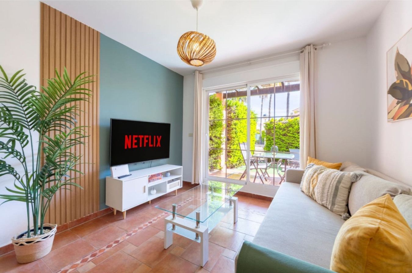 Un salon avec un téléviseur à écran plat affichant le logo « NETFLIX », une table basse en verre, un canapé avec des coussins, une plante en pot et une vue sur l’extérieur grâce à une porte-fenêtre coulissante.