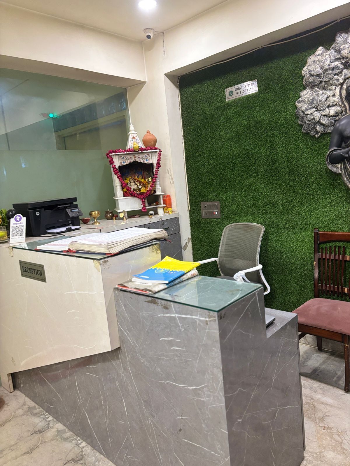 Una reception con una scrivania, un computer, una stampante e un piccolo altare. Si nota una sedia e una parete verde con una statua.