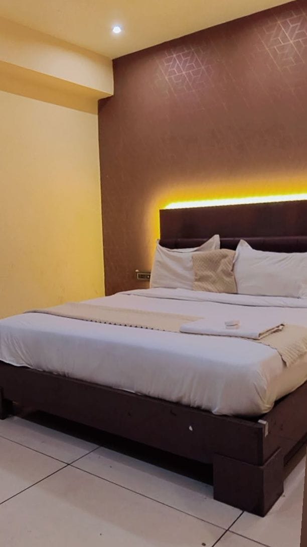 A neatly made bed with white linens and a bedside table, a brown headboard with a geometric pattern, and a tiled floor.