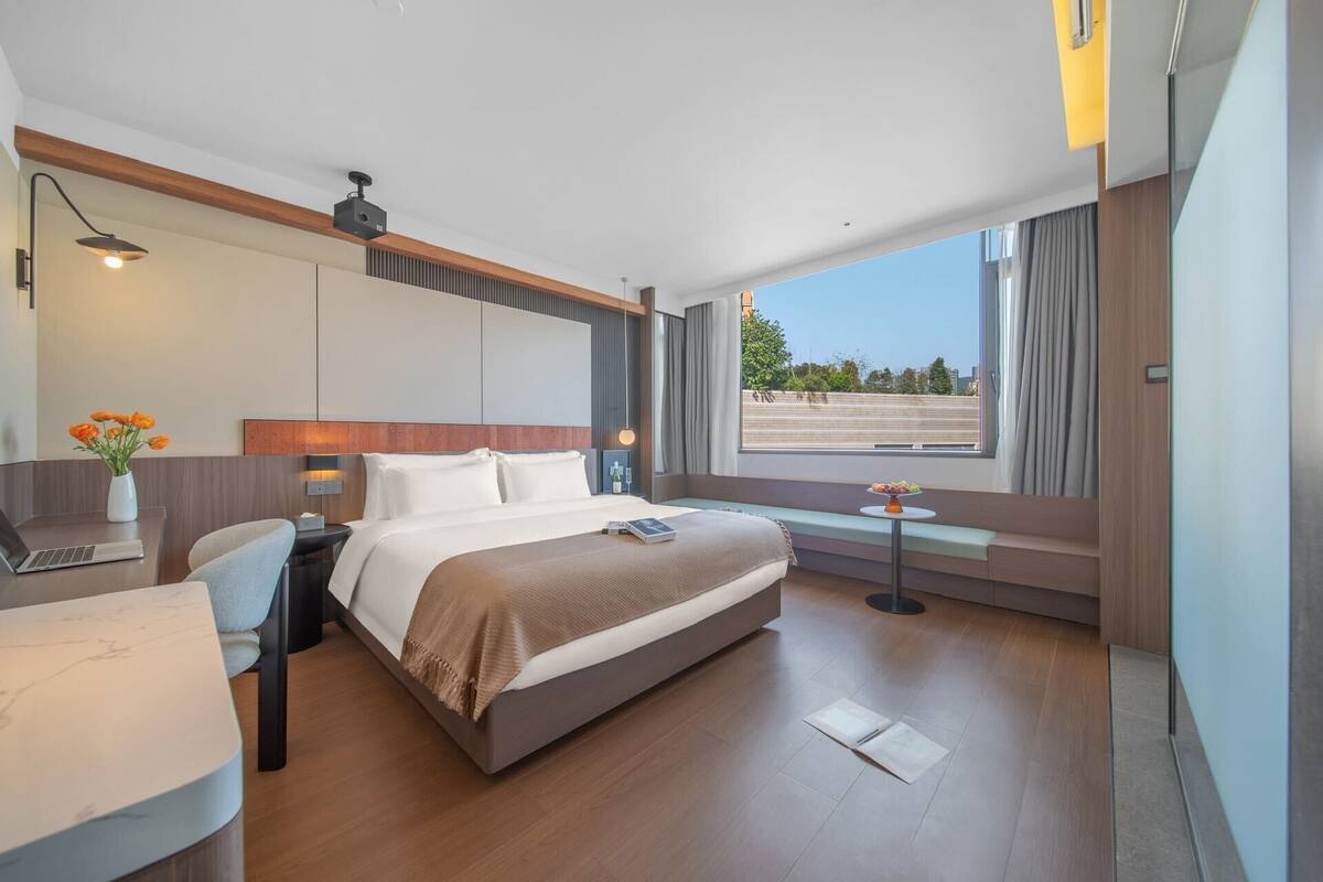 A modern hotel room with a large bed, a desk with a laptop, a small table with flowers, and a view of the city through large windows.