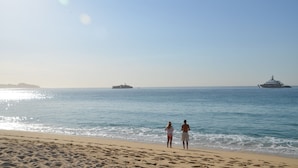 A beach with two people standing by the water, a ship in the distance, and a clear sky.