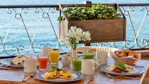 A breakfast table with various dishes, drinks, and a flower vase by a railing with a view of the sea.