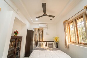 A bedroom with a bed, a ceiling fan, an air conditioning unit, a window with curtains, and a nightstand with a vase of flowers. - Blue Horizon 2bhk Nr Marina Beach Mylapore (Chennai)