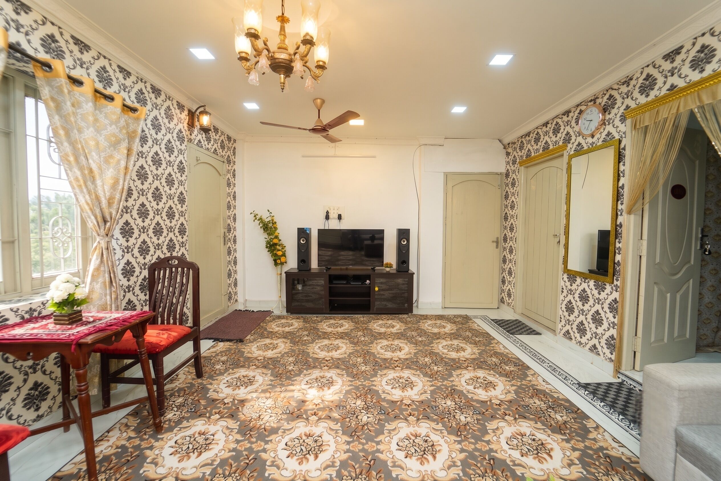 A room with patterned wallpaper, a chandelier, a ceiling fan, a television, and a mirror.