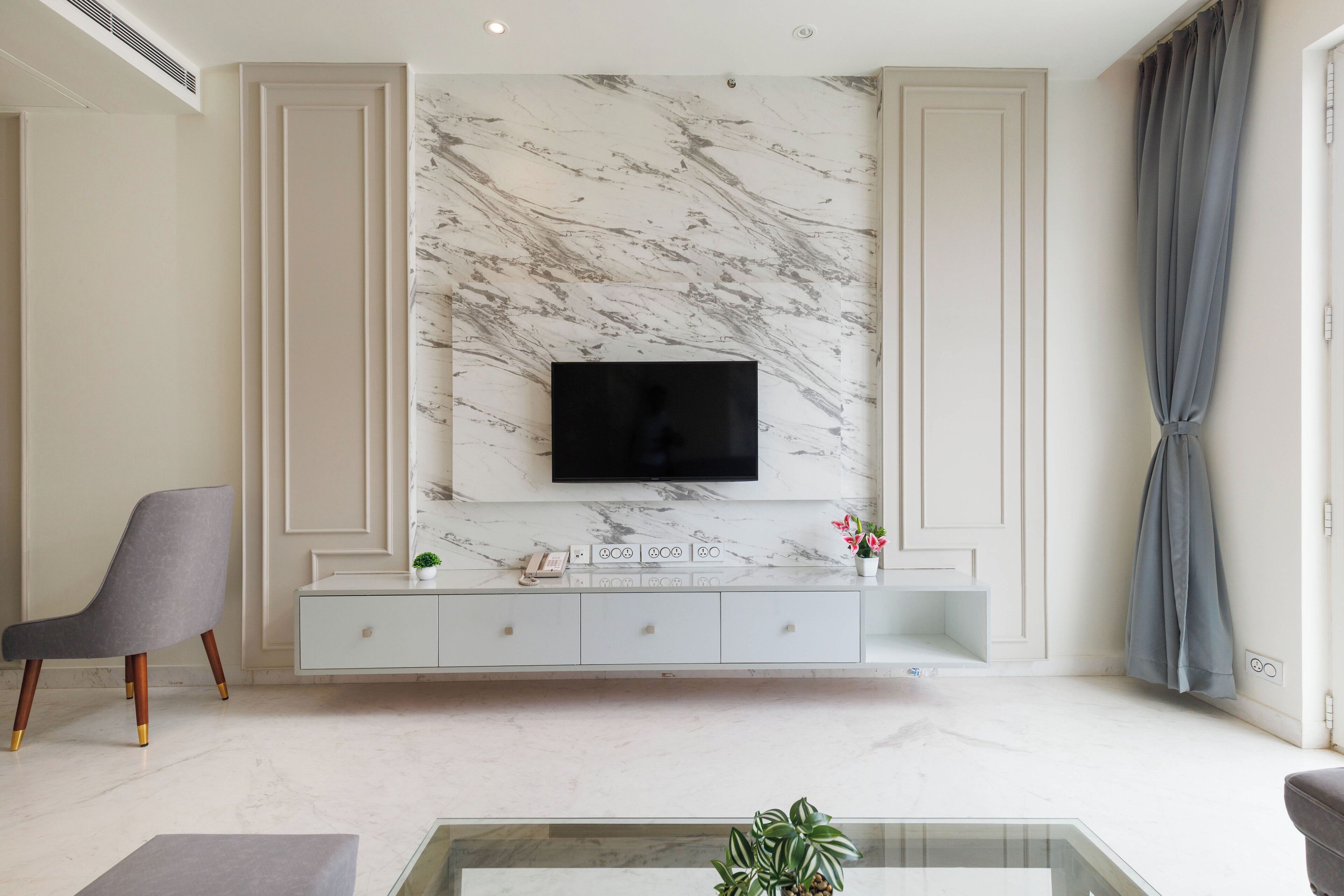 A modern living room with a marble wall, a flat-screen TV, a grey armchair, a glass coffee table, and a white console with decorative items.