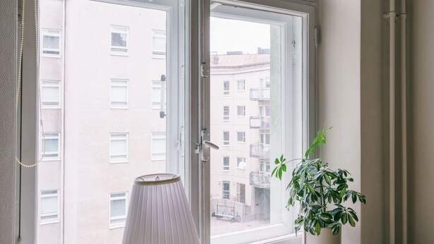 Una habitación con una mesa de madera, una lámpara y una planta en maceta junto a una ventana con vistas a edificios.