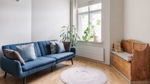 Una sala moderna con un sofá azul, un banco de madera, un amplio ventanal con plantas y un radiador.