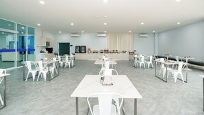 A spacious room with white tables and chairs arranged in rows, a counter with various items, and a glass wall on one side. - One Budget Hotel Chiangrai Maesai Payang (Mae Sai)