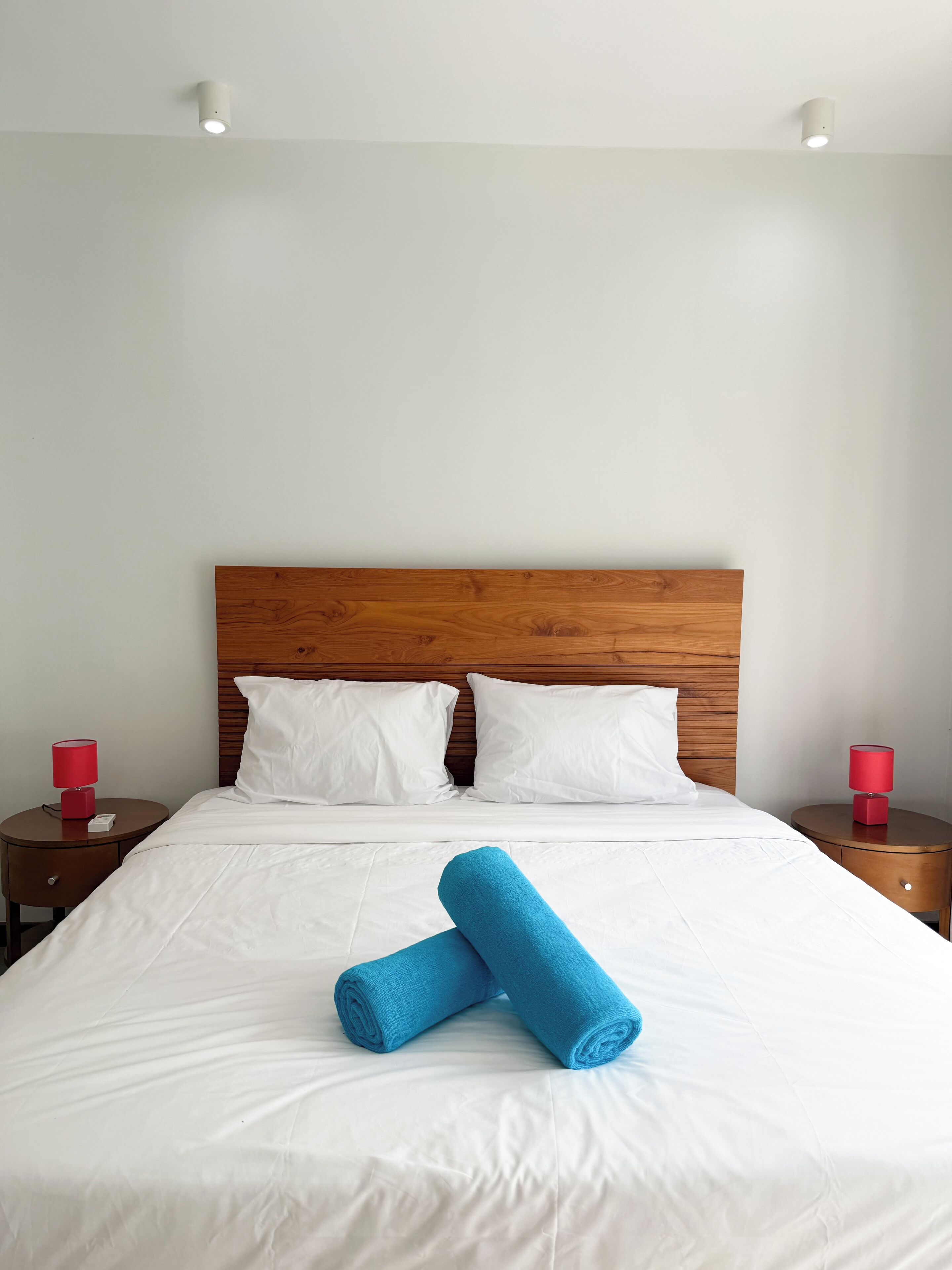 A neatly made bed with white linens and two rolled blue towels, a wooden headboard, and two bedside tables with red lamps.