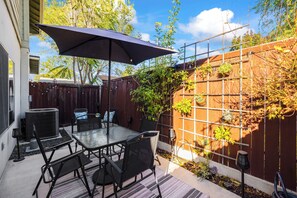 Un patio al aire libre con una mesa y sillas de vidrio, un paraguas grande y una valla de madera con plantas trepadoras.