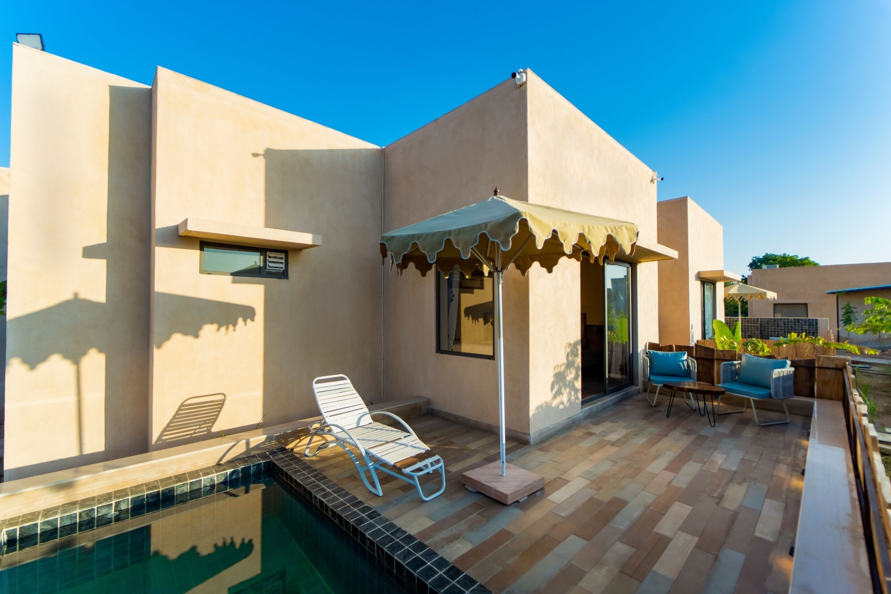 A modern outdoor pool area with a lounge chair, a small table, and a covered seating area with a table and chairs.
