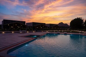 Una piscina con sillas de descanso y un edificio con luces al fondo durante el crepúsculo.