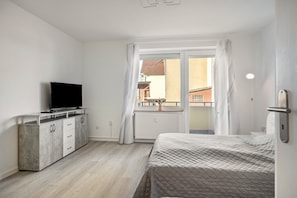 Un dormitorio con una cama, un televisor sobre un mueble, una ventana con cortinas y vistas a edificios.