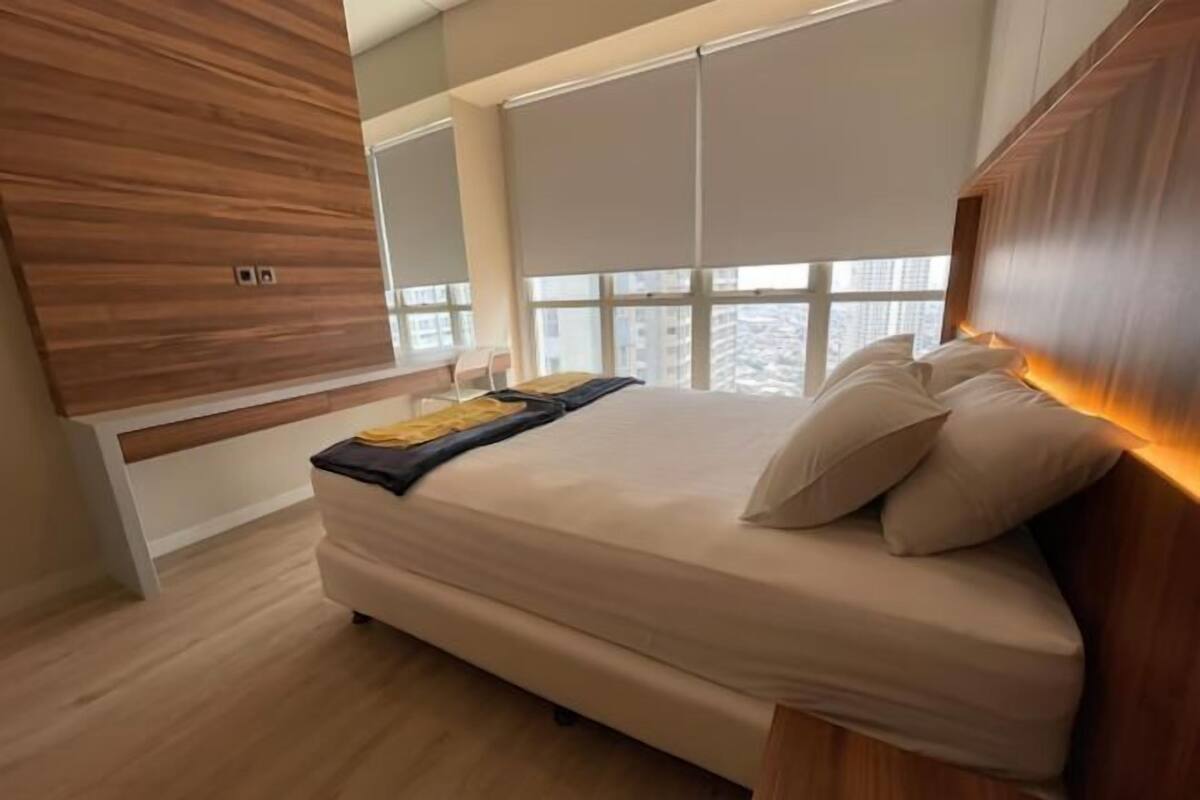 A bedroom with a bed, wooden headboard, large windows with blinds, and a view of the city.