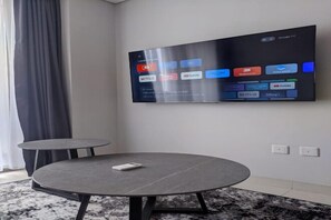 A modern hotel room with a large flat-screen TV mounted on the wall displaying various streaming services. There is a round table with a remote control on it, and two smaller tables. - Premium Taman Anggrek Residences (West Jakarta)