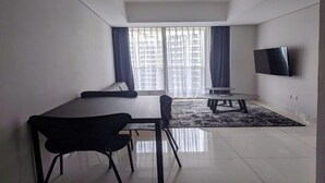 A modern living room with a dining table, chairs, a sofa, a TV mounted on the wall, and a large window with curtains. - Premium Taman Anggrek Residences (West Jakarta)