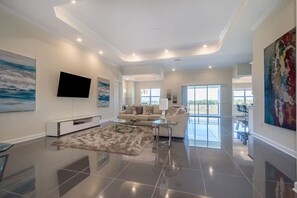 A spacious living room with a large flat-screen TV, a sofa, a coffee table, and a rug. There are large windows and a balcony visible. - Spacious Villa w Pool, Slides & Lush Nature Views (Kissimmee)