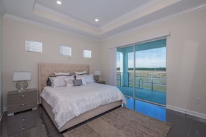 A bedroom with a large bed, bedside lamps, a nightstand, and a view of a blue sky and landscape through a balcony door. - Spacious Villa w Pool, Slides & Lush Nature Views (Kissimmee)