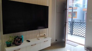 A modern living room with a large flat-screen TV mounted on the wall, a white entertainment unit with drawers, and a sliding glass door leading to an outdoor balcony. - 2D-Villastay (Kovalam)