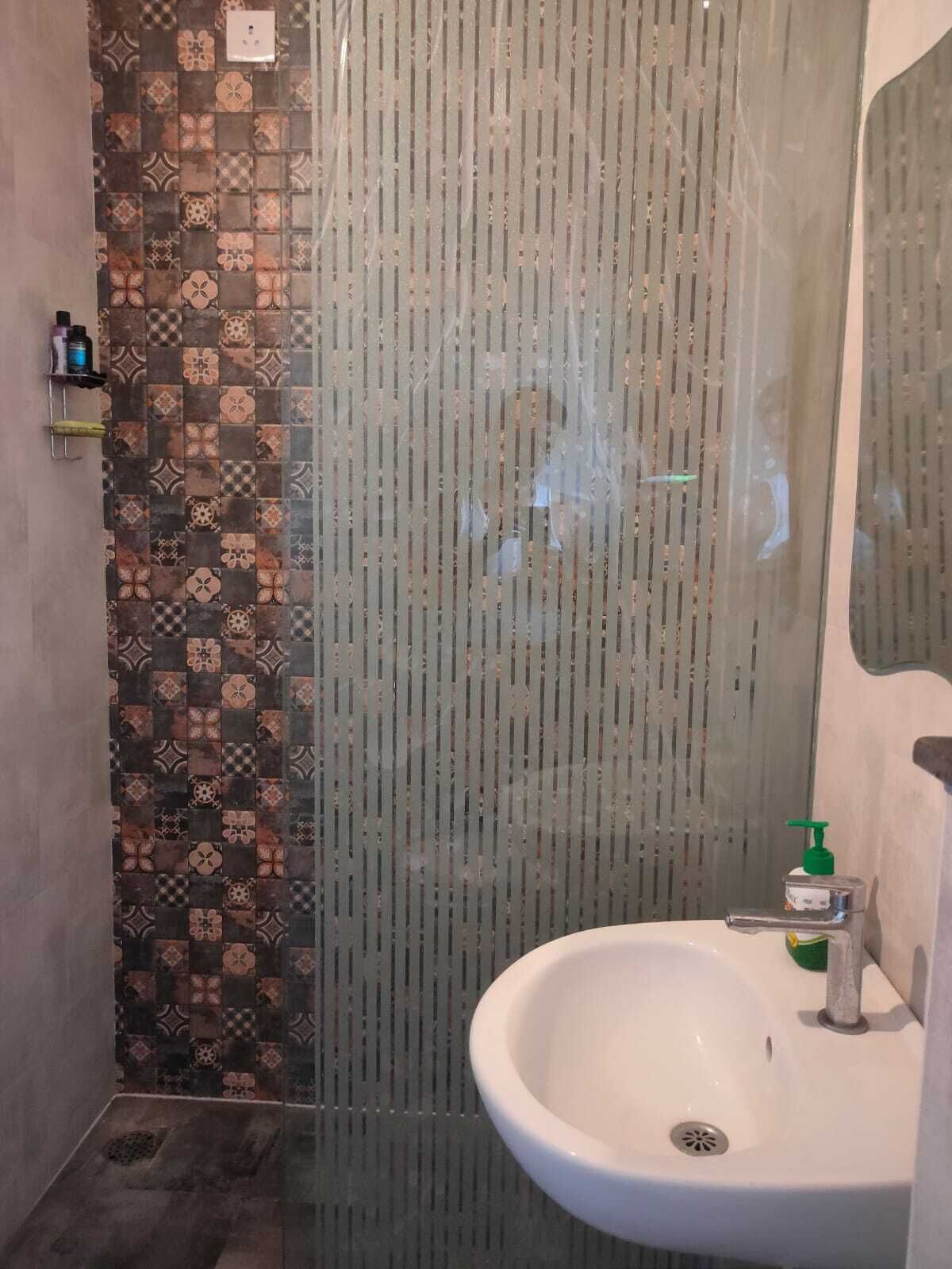 Une salle de bain avec un mur recouvert de carreaux à motifs, une douche en verre et un lavabo blanc agrémenté d’un distributeur de savon vert.