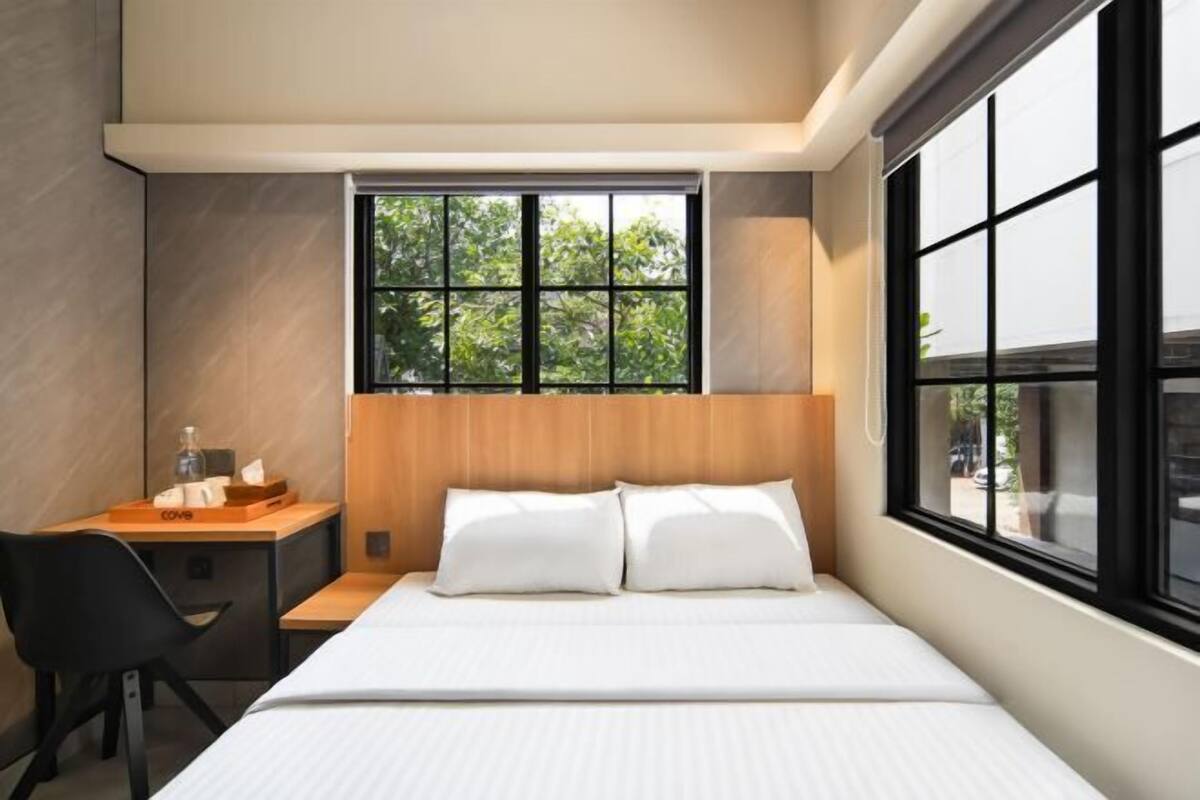 A modern hotel room with a large bed, a wooden headboard, a desk with a chair, and a window with a view of greenery.