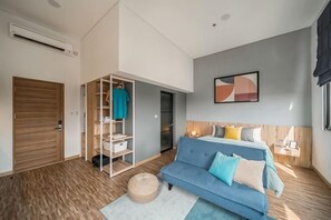 A modern bedroom with a blue sofa, a wooden wardrobe, a wall-mounted abstract painting, and a small bedside table with a lamp. - Cove Prima (South Jakarta)