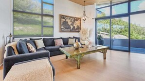 Ein modernes Wohnzimmer mit einer großen Eckcouch, einem Holztisch und großen Fenstern mit Blick ins Freie.