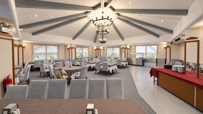 A spacious dining area with multiple tables and chairs, a central chandelier, and large windows providing natural light.