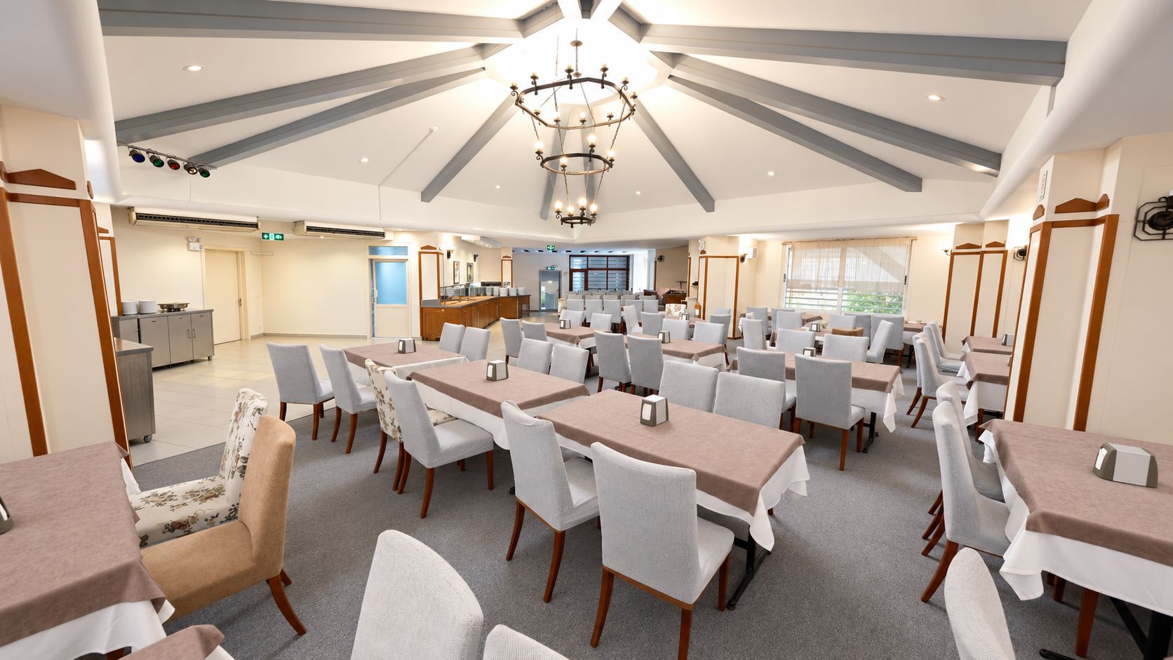 A hotel dining area with multiple tables and chairs, a chandelier, and a kitchen in the background.