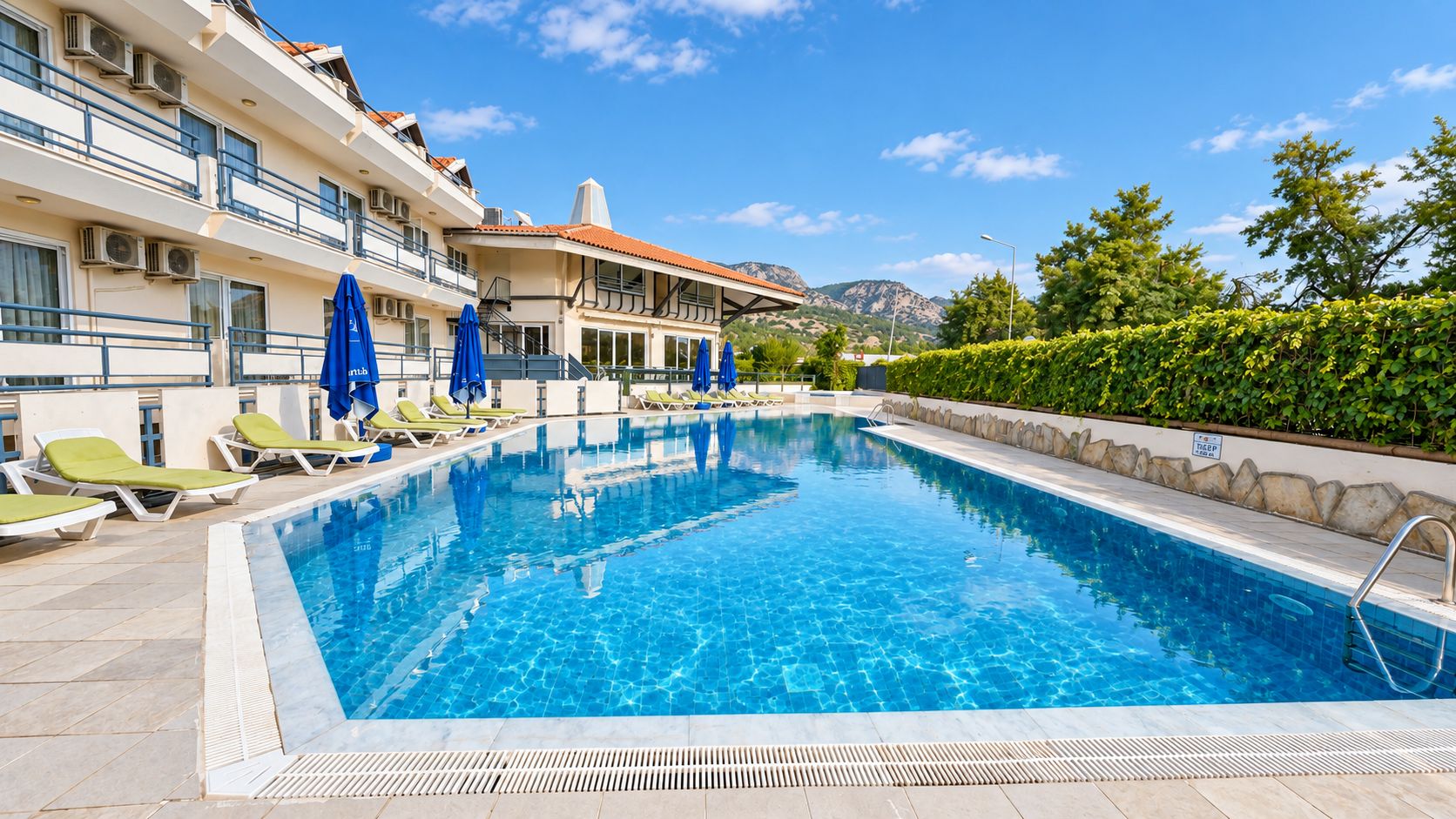 Una piscina con sillones y sombrillas, rodeada por un edificio con balcones y vistas a la montaña.