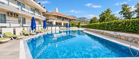 Una piscina con sillones y sombrillas, rodeada por un edificio con balcones y vistas a la montaña.