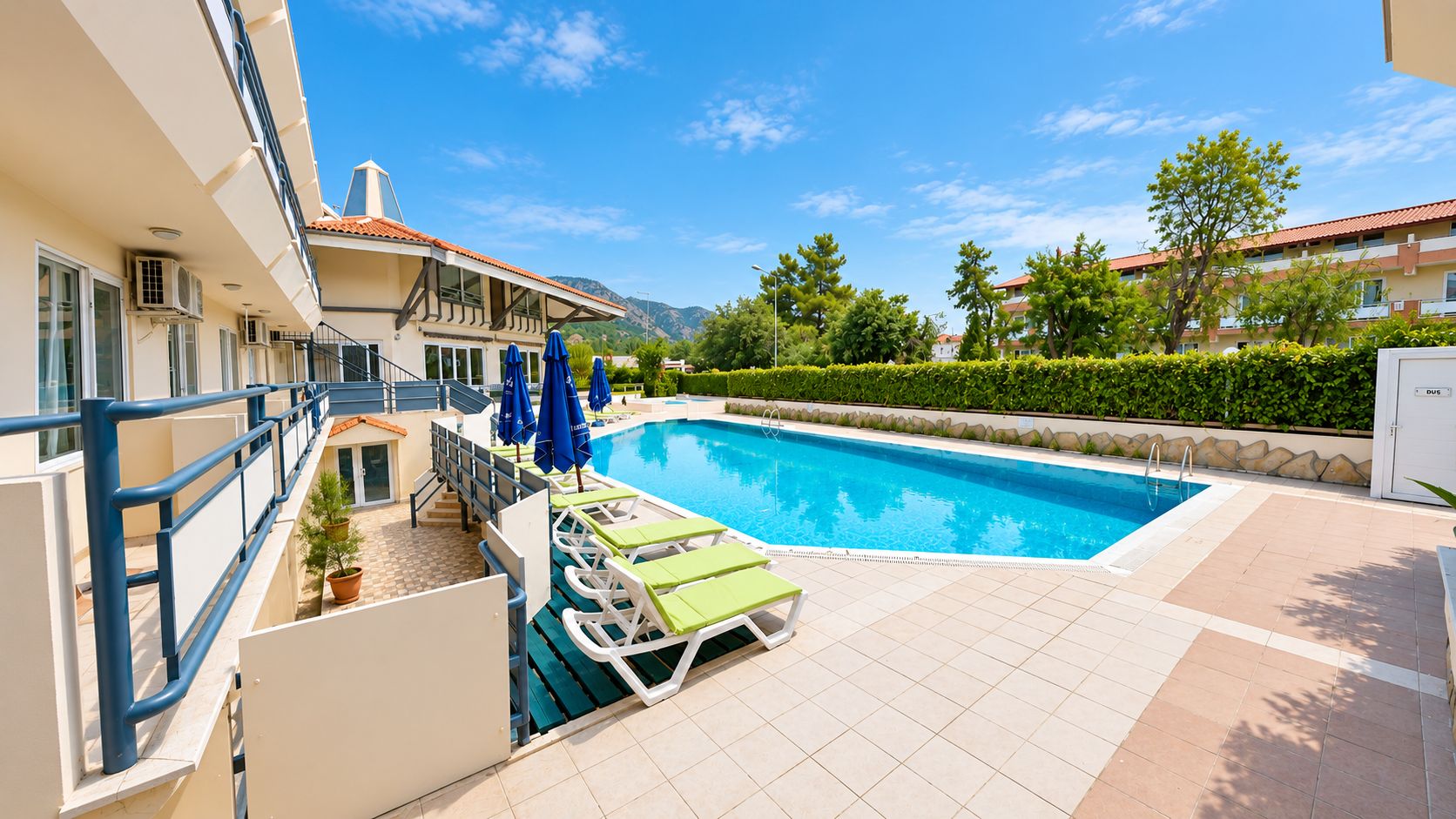 A swimming pool with lounge chairs and umbrellas, surrounded by a building with balconies and a clear sky.