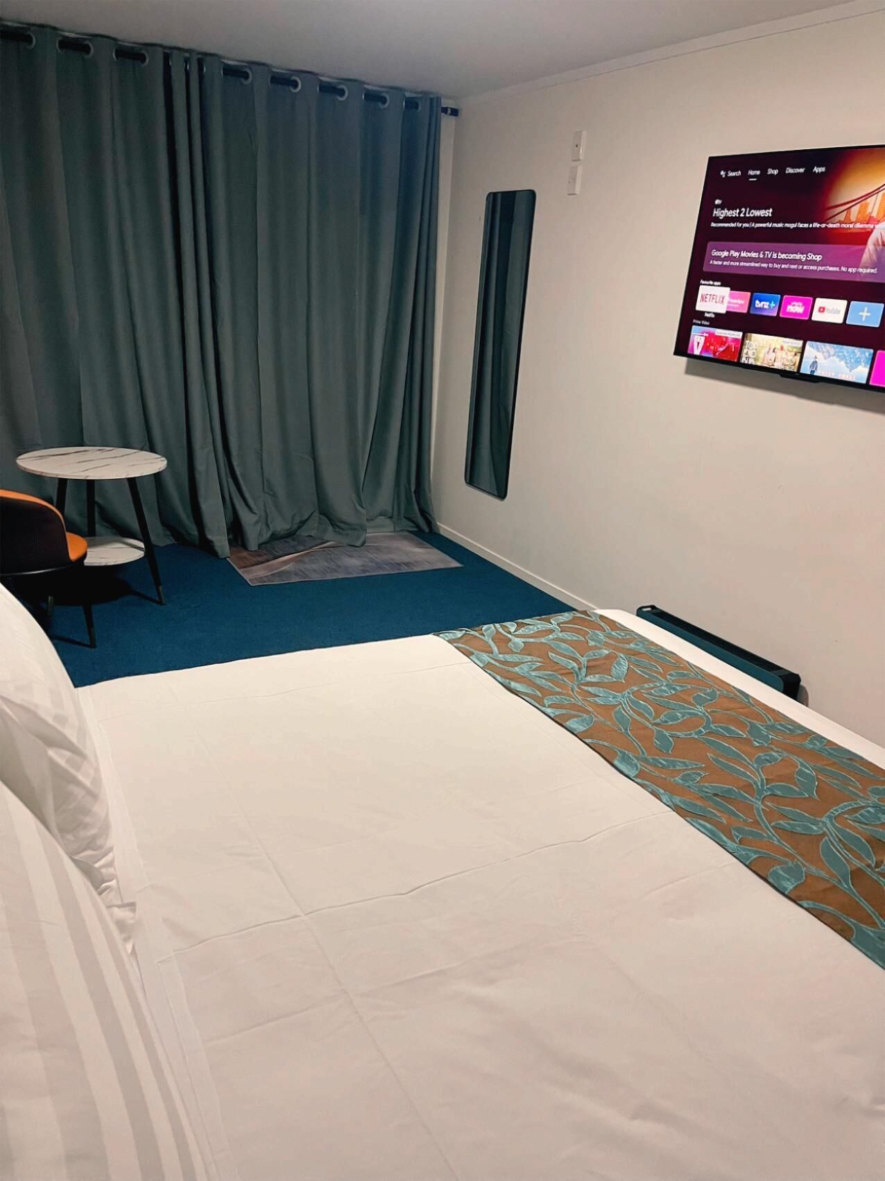 A hotel room with a bed, a small table, a chair, and a TV displaying a movie selection.