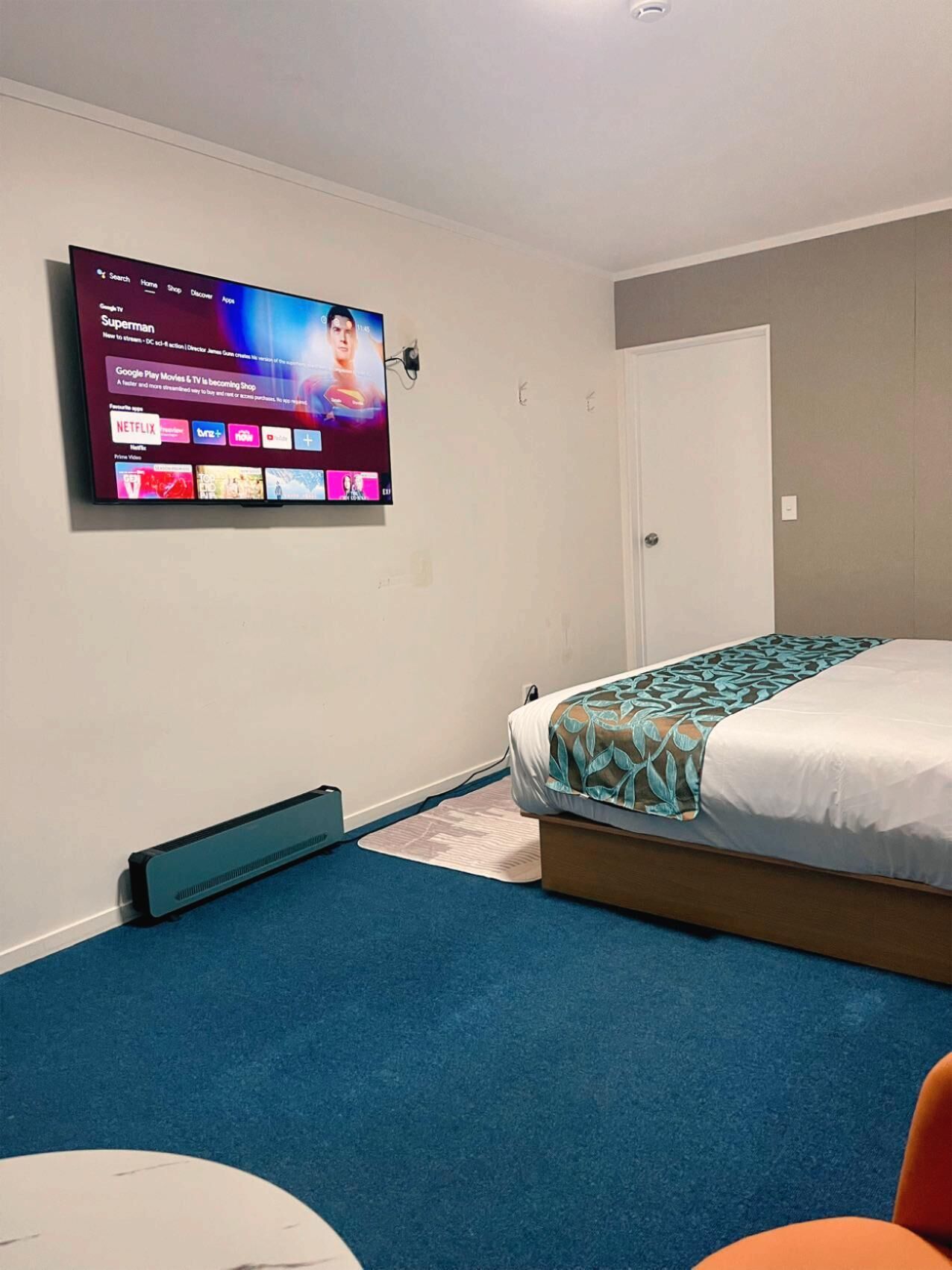 A hotel room with a bed, a wall-mounted TV displaying a streaming service interface, a heater, and a door.