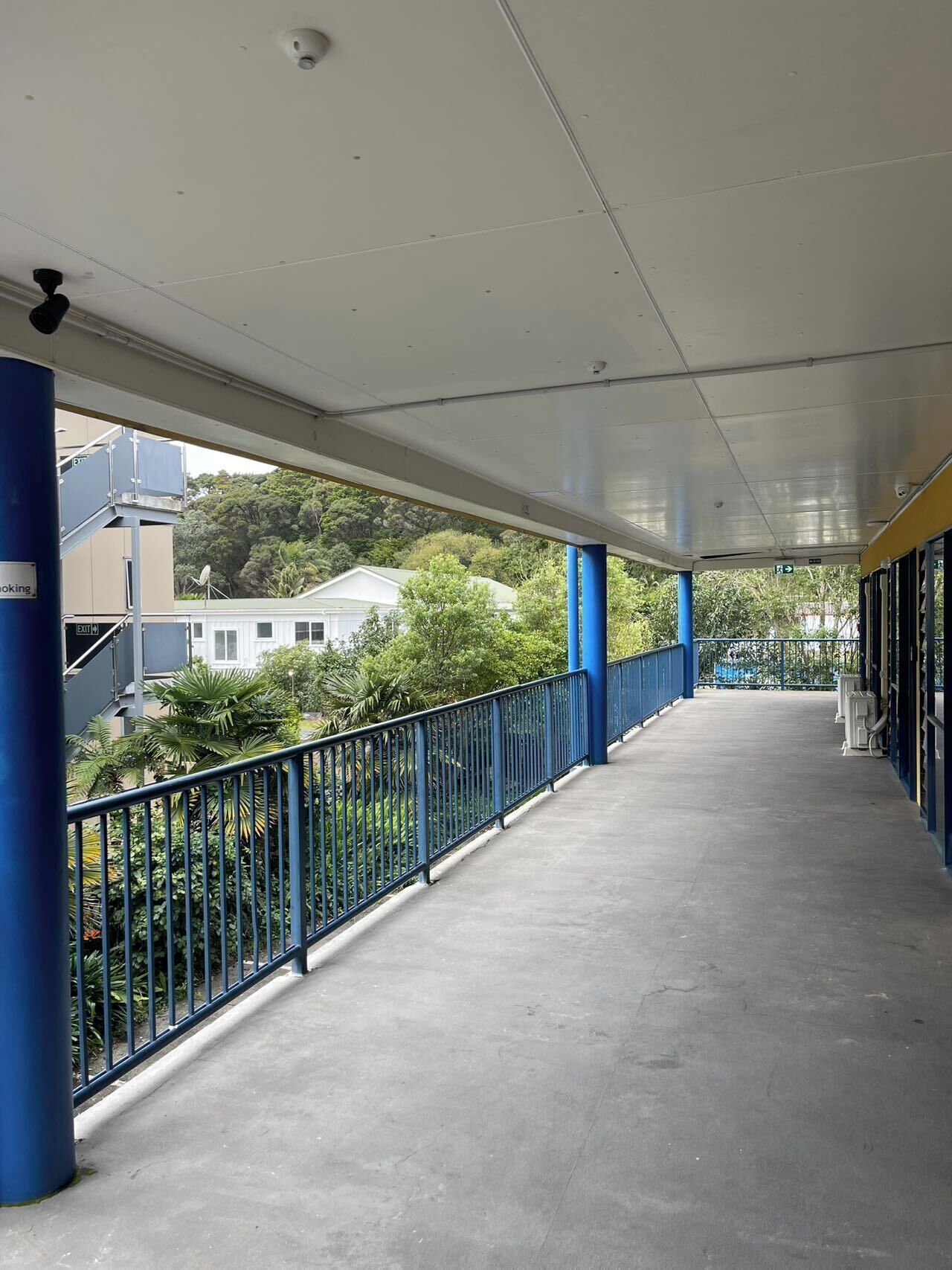 A covered walkway with blue railings, a concrete floor, and a view of greenery and buildings in the background.