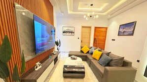 A modern living room with a flat-screen TV, a sectional sofa with yellow and gray cushions, a coffee table, and a wooden accent wall. - Snowdrop Apartments  (Lekki)