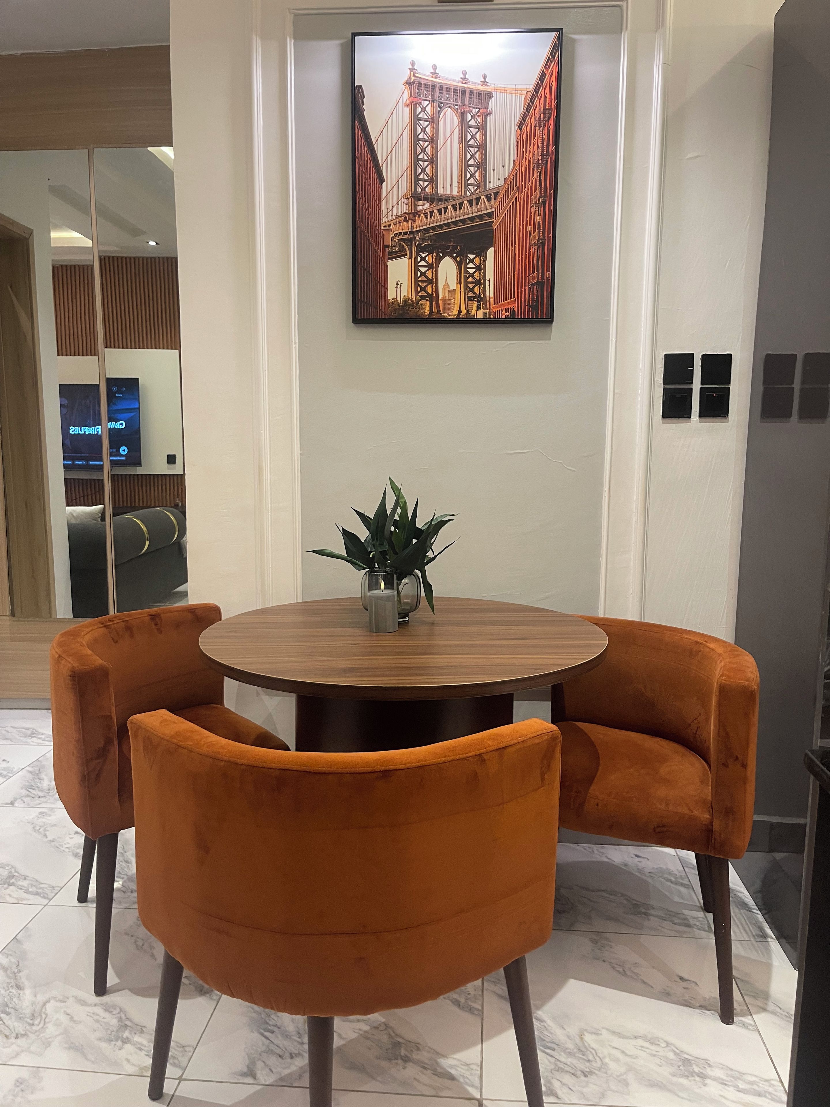 A dining area with a round table, two orange chairs, a potted plant, and a framed picture of a bridge on the wall.