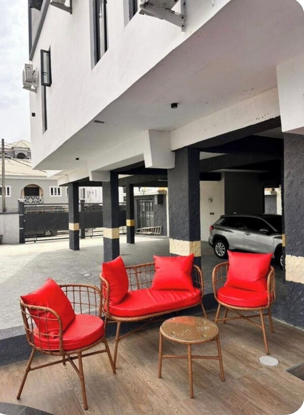 A covered outdoor seating area with wicker chairs and red cushions, a parking garage entrance, and a car parked inside. - Snowdrop Apartments  (Lekki)