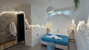A bedroom with a stone wall, a bed with a blue bedspread, a round stool, and a hanging plant. - Ilio Boutique Swim Up Rooms (Ksamil)