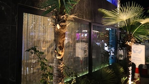 A modern building facade with a large window reflecting city lights, palm trees, and a sign indicating hotel parking. - Ilio Boutique Swim Up Rooms (Ksamil)