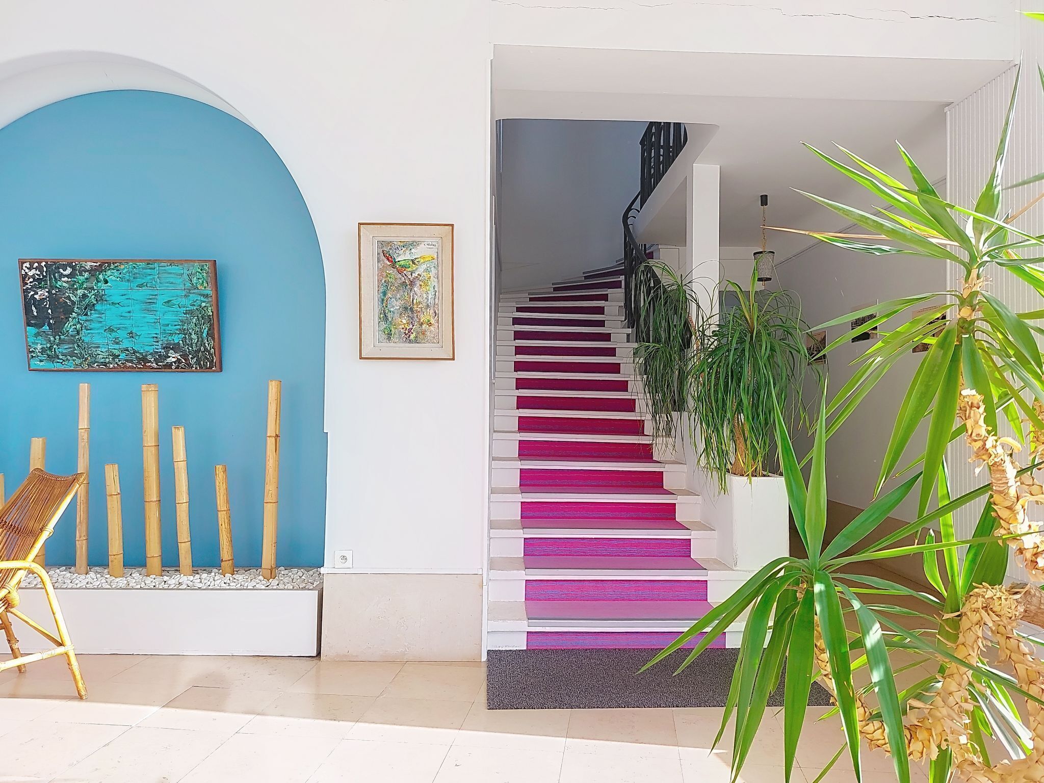 Un escalier aux marches roses menant à un palier, un mur bleu orné d’un tableau encadré et une plante de bambou dans un pot blanc.