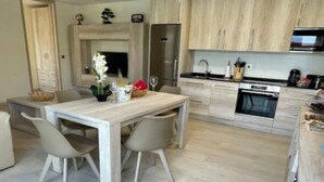 Eine moderne Küche mit hellen Holzschränken, einem Edelstahlkühlschrank und einem eingebauten Backofen. Ein Esstisch mit zwei Stühlen und eine Vase mit Blumen sind ebenfalls vorhanden.