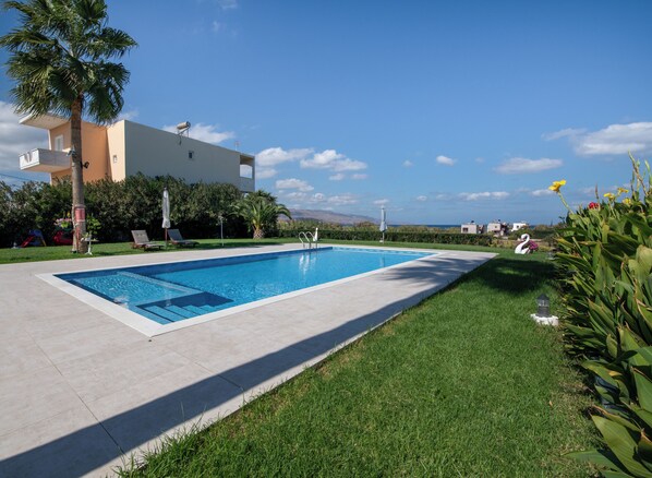 Una piscina con agua cristalina azul, rodeada de un césped cuidado y un edificio de fachada blanca.