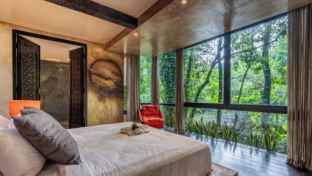A bedroom with a large bed, a red chair, and a view of lush greenery through large windows. - Yara Wellness (Buwit)