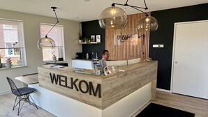 Un mostrador de recepción con letrero 'WELKOM', una lámpara colgante moderna, un panel de madera decorativo y una puerta a la derecha.